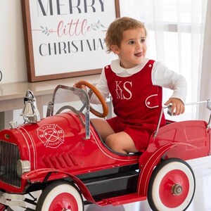 May include: A child in a red outfit with white collar and monogram sits in a vintage red fire truck toy. The toy truck has white-walled tires and a wooden steering wheel. A sign in the background reads "Merry little Christmas."