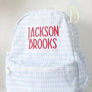 May include: A light blue and white gingham backpack featuring the name JACKSON BROOKS embroidered in red thread on the front. The backpack includes a front zippered pocket with a white zipper pull and side pockets with elasticized openings.