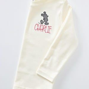 May include: A long-sleeve cream-colored shirt featuring an embroidered black silhouette of a cartoon mouse character positioned above the name CHARLIE stitched in red capital letters on the upper chest area of the garment.