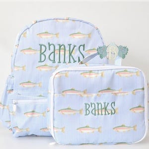May include: A light blue and white pinstriped backpack and matching lunch bag set featuring a repeating trout fish pattern. Both items are embroidered with the name Banks in green thread. A small decorative tag with the letter B is attached.