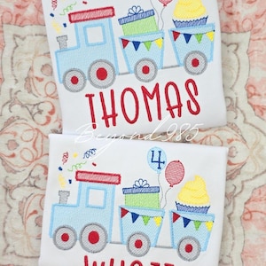 May include: Two white shirts with embroidered birthday train designs. Each train has a blue engine, gift boxes, a cupcake, and balloons. One shirt says "THOMAS" and the other says "WYATT" in red.