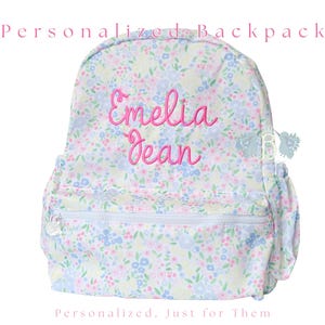 May include: A backpack with a light-colored floral pattern in shades of blue, pink, and green. The name Emelia Jean is embroidered in pink script on the front. The bag features a zippered front pocket, side pockets, and the text Personalized Backpack.