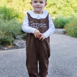 May include: A young child wearing a brown overall with a white collar and a white long-sleeved shirt. The overall has the name "Grayson" embroidered on the front in white thread.