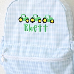 May include: Light blue gingham backpack with a white zipper and a front pocket. The backpack features three green tractors with yellow wheels embroidered above the name "Rhett" in green.