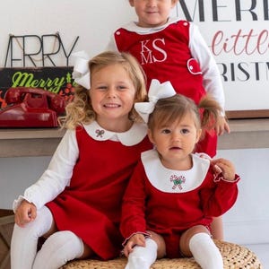 May include: Three children wearing matching red and white Christmas outfits. The outfits include red dresses and rompers with white collars, white bows, and knee-high socks. The outfits have candy cane embroidery. A sign says "Merry Christmas".