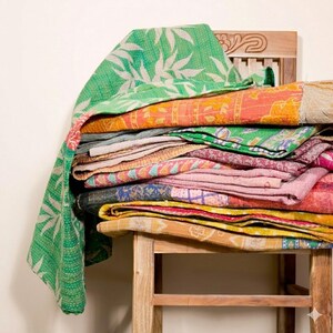 Könnte beinhalten: Ein Stapel gefalteter Kantha-Steppdecken mit verschiedenen Mustern und Farben, darunter Grün mit weißen Blattdrucken, Orange mit Blumenmotiven sowie rosa, gelbe und blaue geometrische Designs, alle mit sichtbaren Steppstichdetails versehen.