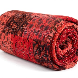 May include: A rolled quilt featuring a patchwork of red and dark brown fabric panels with various floral and abstract patterns. The textile is held together by visible white running stitches that create a textured, quilted appearance throughout.