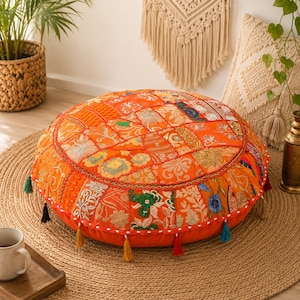 Puede incluir: Un cojín de suelo redondo con un diseño de patchwork en naranja vibrante, bordados florales intrincados, detalles en hilo dorado metálico y pequeñas borlas de tela que cuelgan del borde inferior. La superficie superior consta de varios paneles de tela.