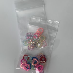 May include: Two clear plastic bags filled with colorful heart-shaped pearl bead rings. The beads are in shades of pink, purple, blue, green, and yellow.