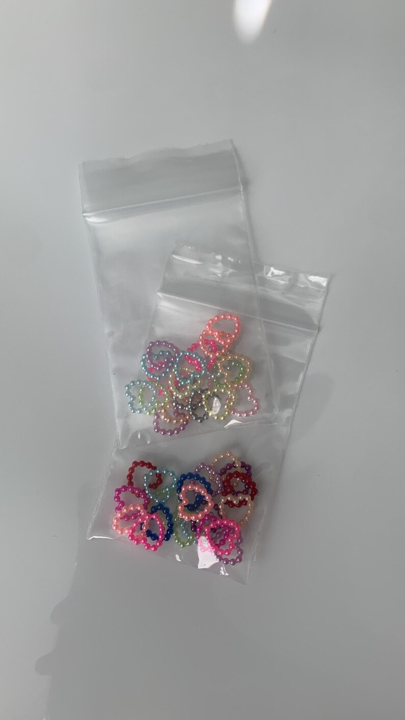 May include: Two clear plastic bags filled with colorful heart-shaped pearl bead rings. The beads are in shades of pink, purple, blue, green, and yellow.