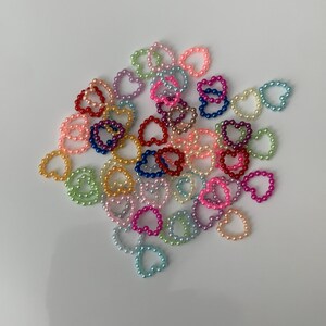 May include: A collection of colorful pearl heart rings. The rings are made of small, round pearls in various colors, including pink, blue, green, yellow, and red. The rings are arranged in a loose pile on a white background.
