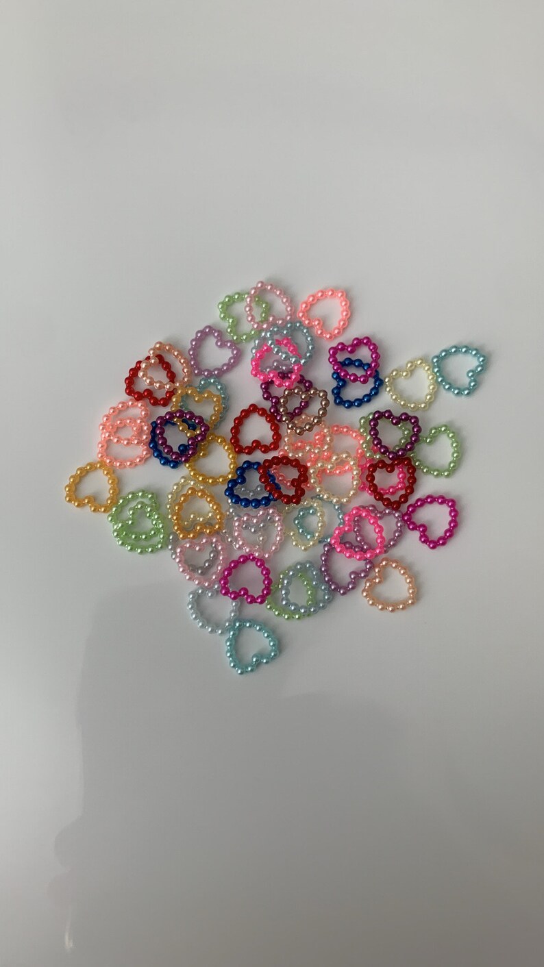 May include: A collection of colorful pearl heart rings. The rings are made of small, round pearls in various colors, including pink, blue, green, yellow, and red. The rings are arranged in a loose pile on a white background.