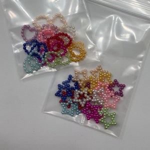May include: Two bags of colorful pearl bead charms in the shapes of hearts and stars. The charms are in a variety of colors, including pink, blue, green, yellow, and white.