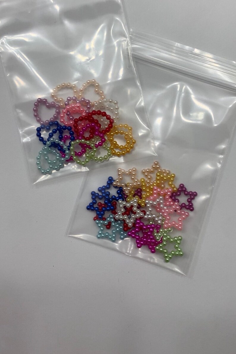 May include: Two bags of colorful pearl bead charms in the shapes of hearts and stars. The charms are in a variety of colors, including pink, blue, green, yellow, and white.