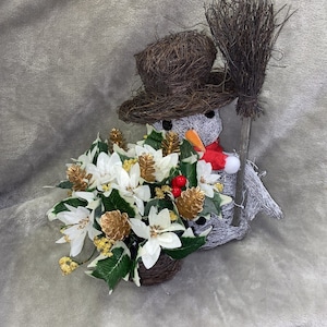 May include: A snowman figurine with a brown twig hat and a red Santa hat, holding a brown twig broom. The snowman is surrounded by a white poinsettia flower arrangement with gold pine cones and green leaves.