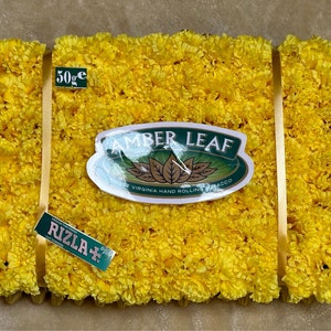 May include: A yellow flower arrangement with a green and white label that says "Amber Leaf Finest Virginia Hand Rolling Tobacco" and a green label that says "Rizla+ Original Rolling Papers". The arrangement is tied with a gold ribbon and has a small white label that says "50g e".