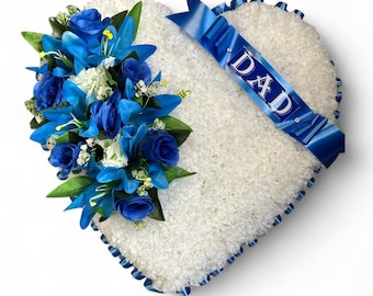 May include: A heart-shaped floral arrangement covered in white flowers with a cluster of blue roses and green leaves on the left side. A blue ribbon diagonally crosses the heart, featuring the white text DAD printed on the fabric.