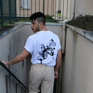 May include: A person wearing a white t-shirt with a black and white graphic print of three figures on the back. The person is wearing beige pants and white sneakers. The person is standing on a set of stairs.