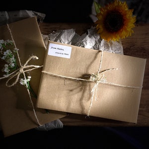 May include: Two brown paper packages tied with twine and decorated with white flowers. The packages are sitting on a wooden surface with a sunflower and crumpled newspaper in the background. One package has a label that reads "From Natura, 2019.02.10 Paris."