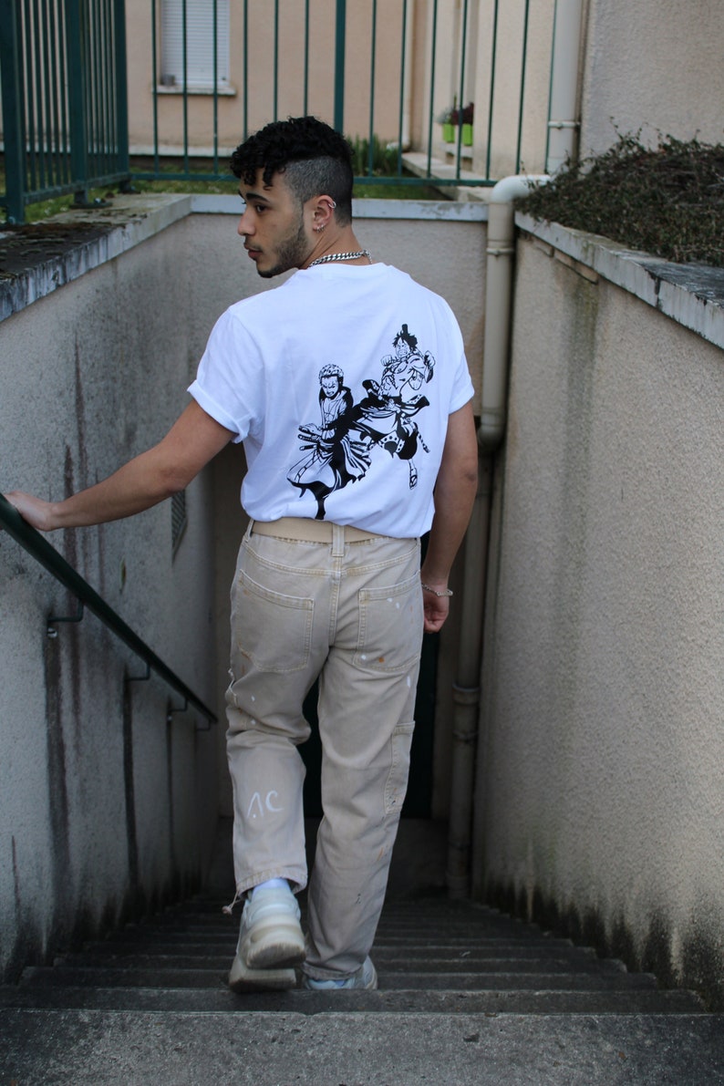 May include: A person wearing a white t-shirt with a black and white graphic print of three figures on the back. The person is wearing beige pants and white sneakers. The person is standing on a set of stairs.