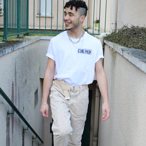 May include: A person wearing a white t-shirt with the text "ONE PIECE" printed on it. They are also wearing beige pants with paint splatters and white sneakers.
