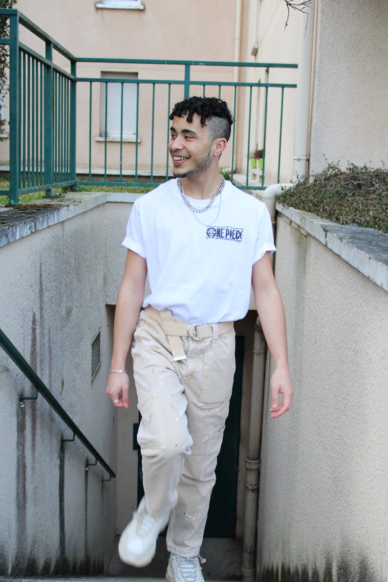 May include: A person wearing a white t-shirt with the text "ONE PIECE" printed on it. They are also wearing beige pants with paint splatters and white sneakers.