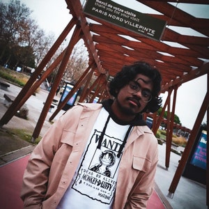 May include: A person wearing a white graphic tee with a "Wanted" poster design featuring a cartoon character, paired with a light brown jacket and pants. The background includes a wooden structure and a sign that reads "PARIS NORD VILLEPINTE".