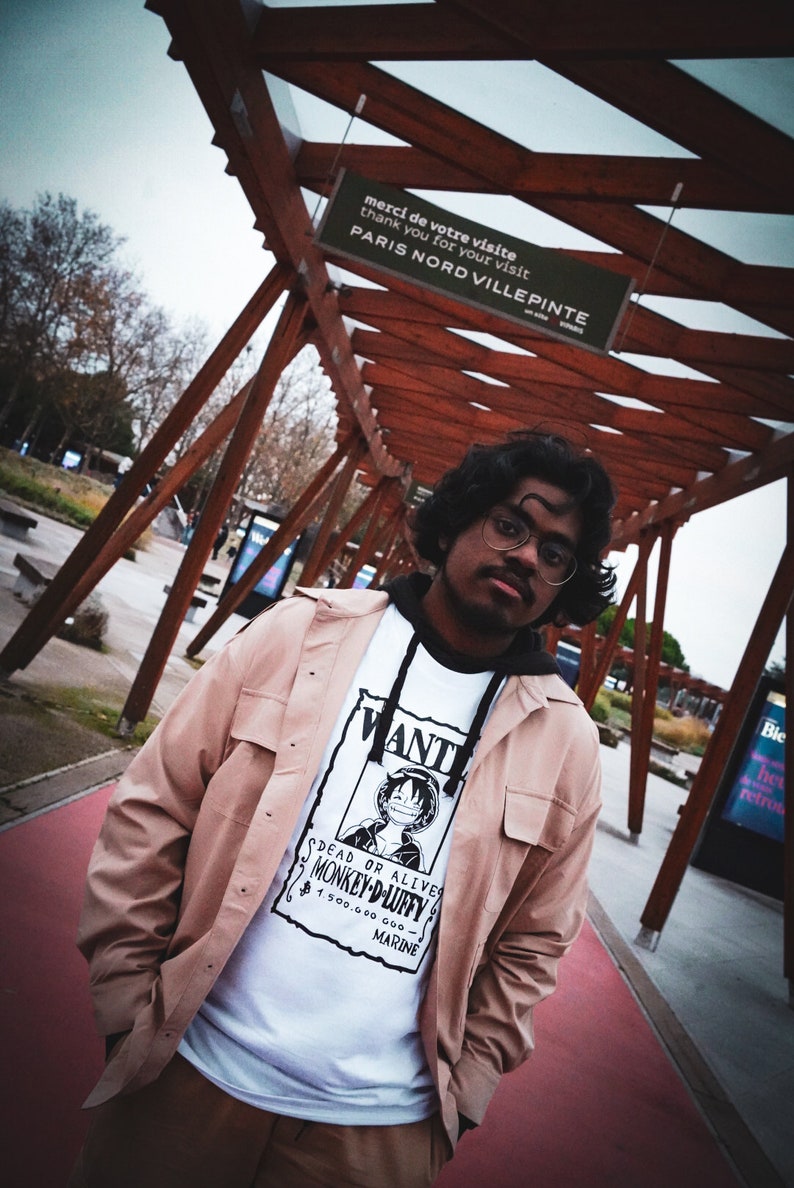 May include: A person wearing a white graphic tee with a "Wanted" poster design featuring a cartoon character, paired with a light brown jacket and pants. The background includes a wooden structure and a sign that reads "PARIS NORD VILLEPINTE".