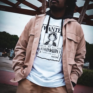 May include: A tan jacket and pants outfit with a white graphic tee featuring a "Wanted" poster of a cartoon character. The poster reads "DEAD OR ALIVE MONKEY D. LUFFY" with a monetary reward. The outfit is worn with a black hoodie.