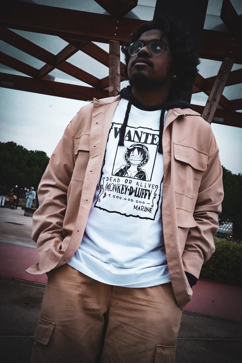May include: A tan jacket and pants outfit with a white graphic tee featuring a "Wanted" poster of a cartoon character. The poster reads "DEAD OR ALIVE MONKEY D. LUFFY" with a monetary reward. The outfit is worn with a black hoodie.