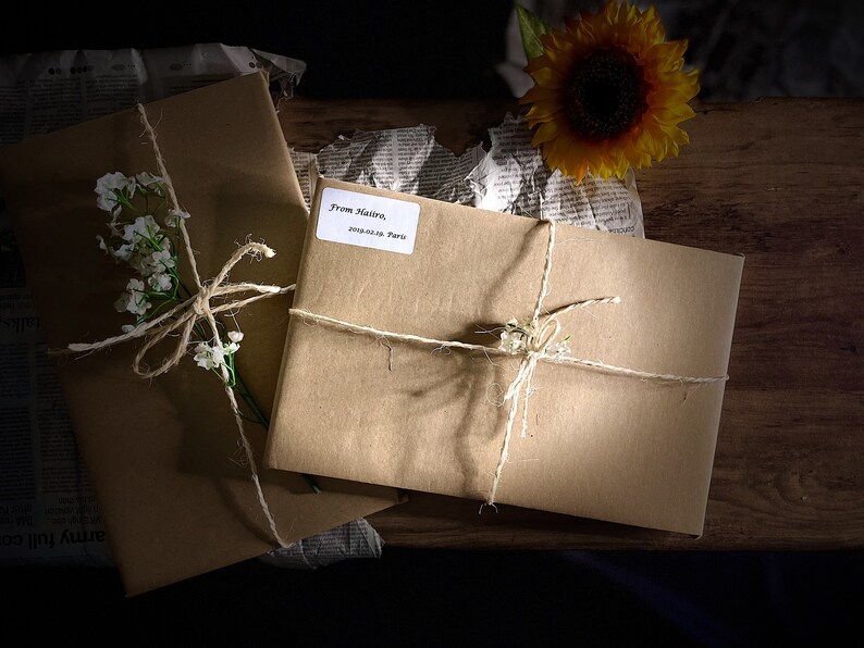 May include: Two brown paper packages tied with twine and decorated with white flowers. The packages are on a wooden surface with a sunflower and crumpled newspaper.