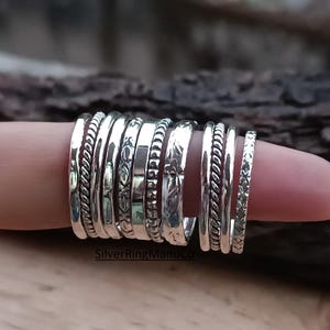 Puede incluir: Un conjunto de anillos de plata con diversas texturas y diseños, incluyendo trenzados, abalorios y grabados. Los anillos están apilados en un dedo, mostrando los diferentes estilos y anchos. La imagen se presenta sobre un fondo de madera borroso.