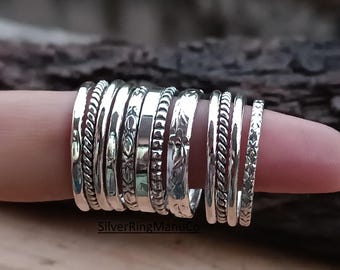 May include: A collection of silver rings with various textures and designs, including twisted rope, beaded, and engraved patterns. The rings are stacked on a finger, showcasing the different styles and widths. The image is set against a blurred wooden background.