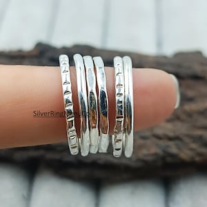 Könnte beinhalten: Eine Sammlung von Silberringen, einige mit strukturierten Designs, wird auf einem Finger präsentiert. Die Ringe variieren im Stil, einige haben eine glatte, polierte Oberfläche, andere weisen gravierte Muster auf. Die Ringe sind gestapelt.