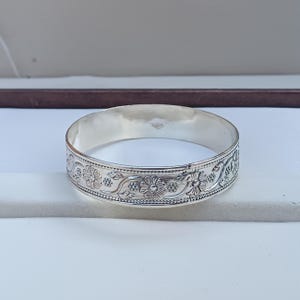 Könnte beinhalten: Ein silbernes Armreif-Armband mit floralem Design. Das Armband ist glatt und glänzend.