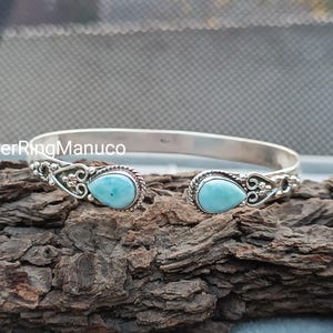 Op de afbeelding: Een zilveren manchetarmband met twee peervormige, lichtblauwe edelstenen. De armband heeft een open ontwerp met decoratieve filigraan details rond de edelstenen. De armband wordt getoond op een stuk getextureerd hout.