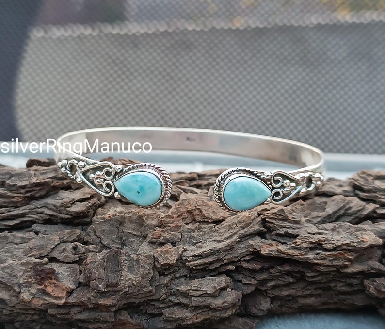 Op de afbeelding: Een zilveren manchetarmband met twee peervormige, lichtblauwe edelstenen. De armband heeft een open ontwerp met decoratieve filigraan details rond de edelstenen. De armband wordt getoond op een stuk getextureerd hout.