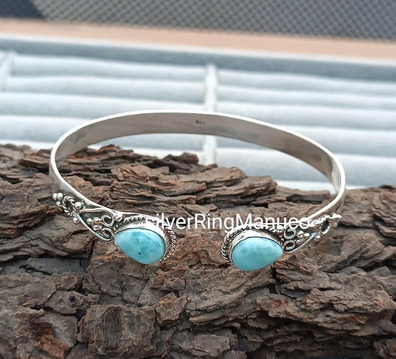 Op de afbeelding: Een zilveren manchetarmband met sierlijke details en twee lichtblauwe, druppelvormige stenen. De armband is aan de onderkant open en ligt op een getextureerd, bruin oppervlak.