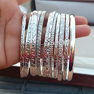 Puede incluir: Un conjunto de pulseras de plata con diseños intrincados. Las pulseras están apiladas una encima de la otra y tienen una variedad de patrones, que incluyen diseños florales, geométricos y con forma de onda.