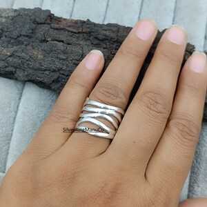 Könnte beinhalten: Ein silberner Ring mit einem geschichteten Design wird auf einer Hand gezeigt. Der Ring hat mehrere horizontale Bänder, die ein strukturiertes, offenes Aussehen erzeugen. Der Ring wird an einem Finger getragen und zeigt seine komplizierten Details und seine Handwerkskunst. Der Ring ist ein Schmuckstück.