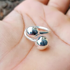 Könnte beinhalten: Ein silberner Ring mit zwei kugelförmigen, reflektierenden Kugeln. Das Band des Rings umschließt die Kugeln und erzeugt ein einzigartiges Design. Der Ring wird auf einer Hand gezeigt.