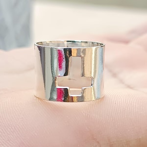 Könnte beinhalten: Silberring mit einem ausgeschnittenen Kreuzdesign. Der Ring ist breit und hat eine polierte Oberfläche.