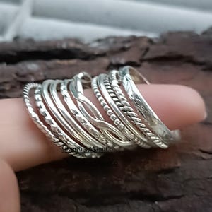 Könnte beinhalten: Eine Sammlung silberner Ringe mit verschiedenen Texturen, darunter gedrehte Seile, Perlen und glatte Bänder. Die Ringe sind auf einem Finger gestapelt und zeigen die verschiedenen Designs und die reflektierende Qualität des Metalls. Die Ringe sind in verschiedenen Größen und Stilen.