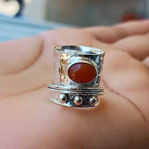 Könnte beinhalten: Ein silberner Ring mit einem ovalen, rot-orangen Stein. Der Ring hat ein strukturiertes Band und einen drehbaren Mittelteil mit drei kleinen, runden Verzierungen. Der Ring wird auf der Handfläche einer Person gezeigt.