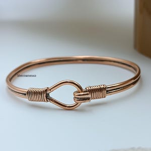 Könnte beinhalten: Ein kupferfarbenes Armreif-Armband mit Hakenverschluss. Das Armband hat ein schlichtes, elegantes Design mit gewickelten Drahtdetails auf jeder Seite des Hakens. Das Armband wird auf einer weißen Oberfläche präsentiert.