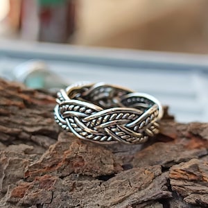 May include: A silver-colored ring with a braided design. The ring features an intricate pattern of interwoven strands, creating a textured surface. The ring is set against a natural, textured background.