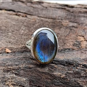 Könnte beinhalten: Ein Silberring mit einem großen, ovalen Labradorit-Edelstein. Der Stein zeigt irisierende blaue und goldene Farbtöne. Der Ring ist auf einer strukturierten, dunkelbraunen Holzoberfläche platziert.