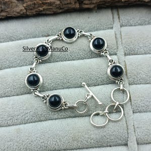 Könnte beinhalten: Silberarmband mit sieben runden, schwarzen Edelsteinen. Das Armband hat einen Knebelverschluss und wird auf einer grauen Oberfläche präsentiert. Der Text "SilverKingManuCo" ist im Bild sichtbar.