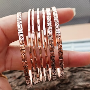 Könnte beinhalten: Ein Satz kupferfarbener Armreifen. Einige Armreifen haben ein Blumenmuster, während andere schlicht sind. Die dünnen Metallarmreifen zeigen ein Blumendesign mit Blättern und Blüten. Die Armreifen sind zusammen gestapelt.