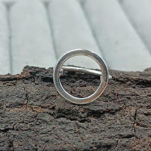 Könnte beinhalten: Ein silberner Ring mit kreisförmigem Design. Der Ring ist vor einem dunklen, strukturierten Hintergrund platziert, der seine schlichte, moderne Ästhetik hervorhebt. Der Ringsteg ist dünn, und der Kreis ist offen, was einen minimalistischen Look erzeugt.